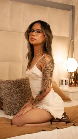 Tattooed woman in a white summer dress sitting on a bed in a warm, cozy bedroom setting.
