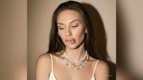 Elegant woman with long brown hair wearing a white tank top and layered pearl necklaces in soft studio lighting.