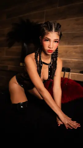 Young woman with braided hair in a black outfit posing in a moody, dimly lit room.