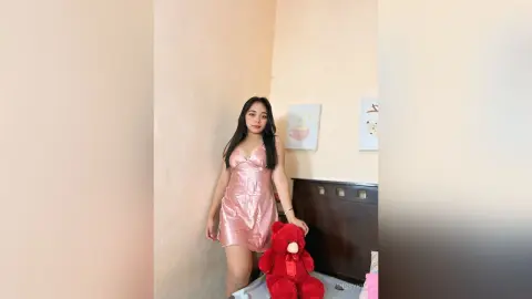 Young woman in a pink satin slip dress sitting in a cozy bedroom next to a large red teddy bear.