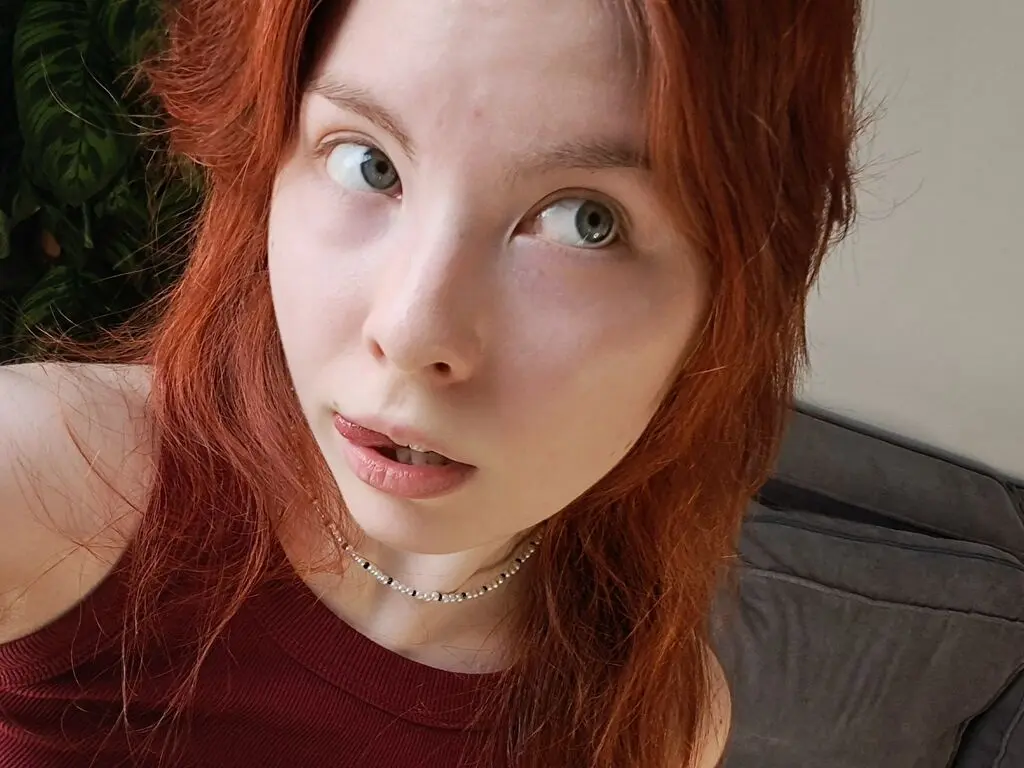 Redheaded woman in a burgundy tank top looking thoughtfully at the camera in a cozy indoor setting.