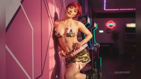 Red-haired woman in a camo bikini and pink mini skirt posing against a pink wall in a neon arcade setting.