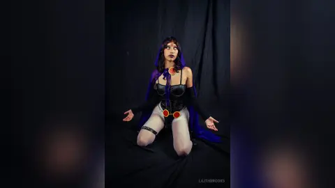 LilithBrooks poses in a black corset and purple hooded cape, wearing white undies with red button details while kneeling on a black surface.