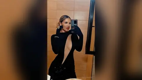 TiffanyAddams poses in a mirror wearing a black long-sleeved dress with a deep, plunging neckline and black gloves.