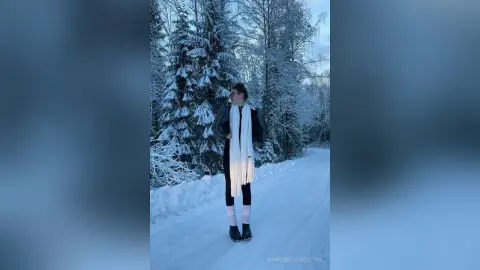 Woman in black winter coat and white scarf standing in a snowy pine forest during a peaceful winter day.