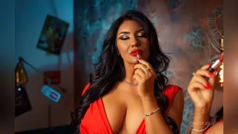 MissyJolie applies red lipstick while wearing a low-cut red dress and a silver necklace.