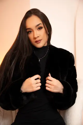 Elegant woman with long dark hair wearing a black turtleneck and faux fur coat in a warm, soft-lit setting.