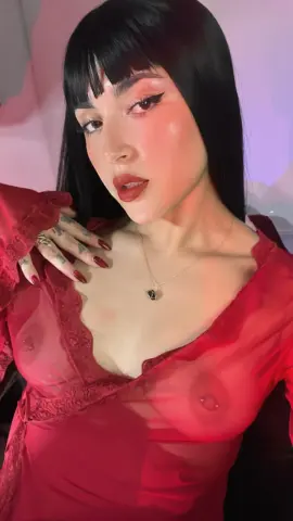 Young woman with black bangs in a sheer red lace top posing in soft pink lighting.