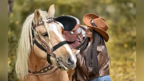 Woman in brown leather jacket petting a white horse in a soft-focus autumn forest setting.