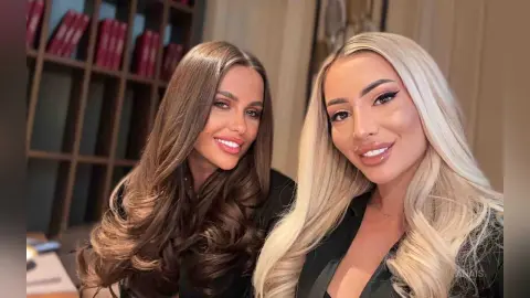 Two smiling women with long wavy hair posing together in elegant black outfits in a cozy indoor setting.