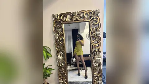 NicoleBella poses in a short yellow dress and black strappy heels, standing before a large, ornate gold mirror.