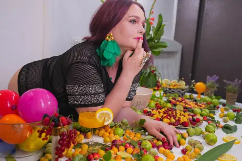 Woman in black mesh top leaning over a colorful spread of fruit and bright balloons in a vibrant setting.
