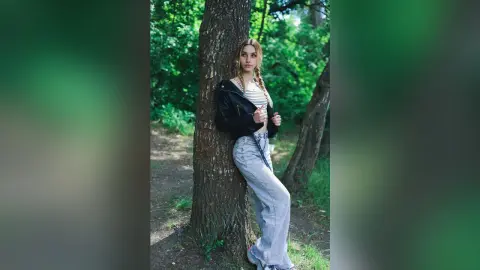 Young woman in a black leather jacket and light wash jeans leaning against a tree in a lush green forest.