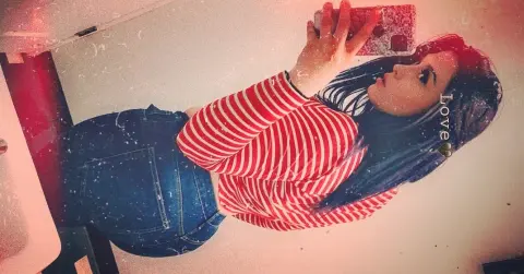 Exoticlexi poses in a red and white striped long-sleeve shirt and blue denim jeans, holding a red phone to take a mirror selfie.