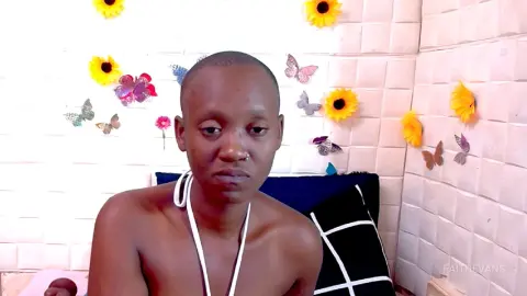 A young man with a shaved head wearing a white halter top in a bright room decorated with sunflower wall decals.