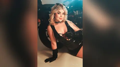 FiorellaRossi poses in a black shiny latex bodysuit and black gloves against a dark motorcycle background.
