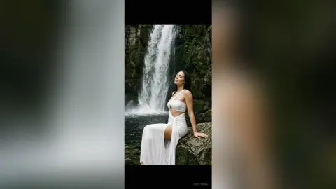 Serene woman in a flowing white maxi dress sitting on a rock by a lush tropical waterfall.