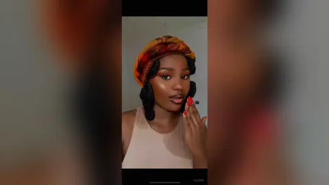 Valjohn wears a patterned headwrap and a tan tank top, posing with her hand near her lips.