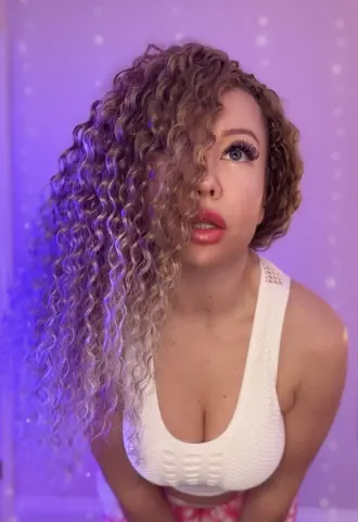 KatBigBum — Young woman with long curly blonde hair wearing a white tank top in soft purple lighting.