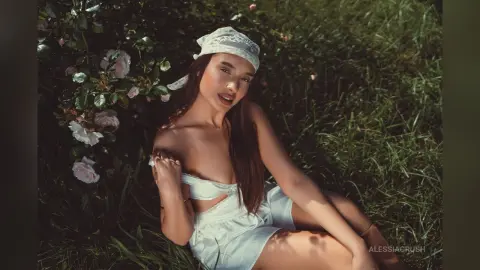 AlessiaCrush sits in the grass by some roses, wearing a white lace headband and a white top that slips off her shoulder.