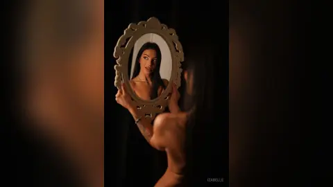 Iza — Young woman with long dark hair looking into an ornate vintage mirror in a moody, dimly lit setting.