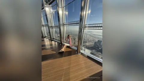 Woman in a white top sitting on a sunny skyscraper balcony overlooking a modern city skyline.