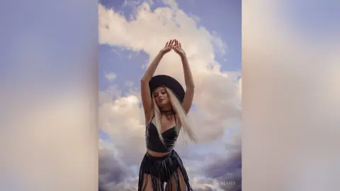 BeaNix poses with her arms raised above her head, wearing a black leather top and a black hat against a cloudy sky.