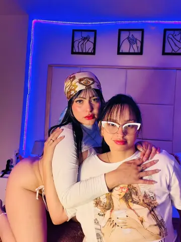 Two young women in white outfits posing together in a bedroom with moody blue neon lighting.