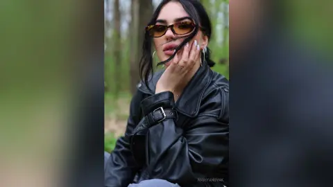 Stylish woman in a black leather jacket and sunglasses posing in a lush green forest setting.