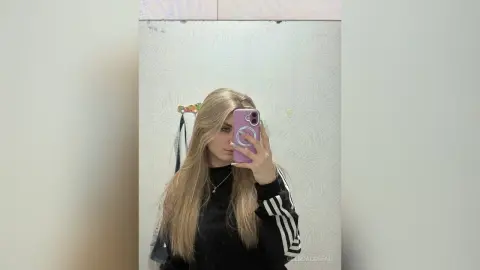 Young woman with long blonde hair in a black sweatshirt taking a moody mirror selfie in a bright room.