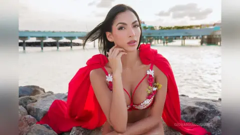 Beautiful woman in a red floral bikini sitting on rocks by the ocean at sunset.