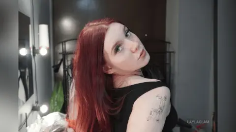 Red-haired woman in a black tank top posing with a moody expression in a modern indoor setting.