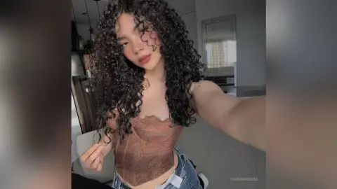 Young woman with long curly hair wearing a brown lace corset top in a modern minimalist room.