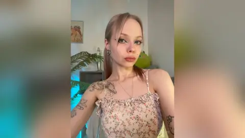 LucyLoy poses in a floral print top, showing off her tattoos and blonde hair in a bright room.