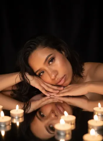 Beautiful woman with wavy hair resting her chin on a reflective surface near glowing candles in a dark setting.