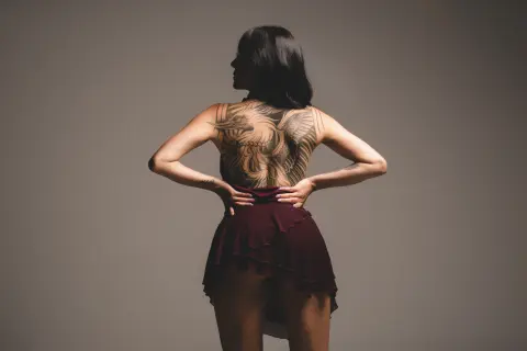 Back view of a woman with large back tattoos wearing a burgundy skirt in a minimalist studio setting.