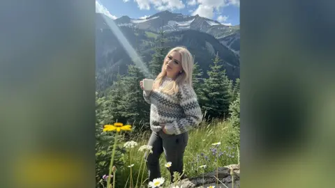 Blonde woman in a cozy patterned sweater holding a coffee mug in a sunny mountain meadow landscape.