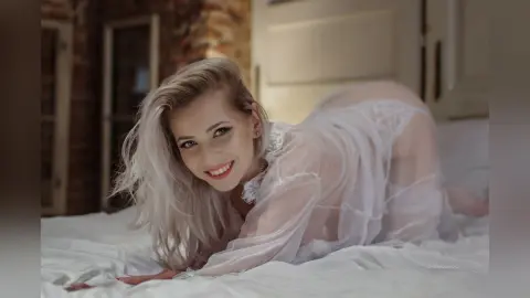 Smiling blonde woman in a sheer white lace outfit lounging on a white bed in a cozy, romantic bedroom setting.