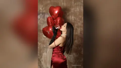Elegant woman in a red satin dress and black gloves drinking from a glass near red heart balloons.