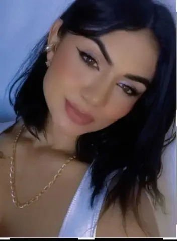 Young woman with dark hair and gold chain necklace posing with soft makeup in a bright setting.