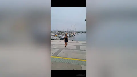 Woman in a black dress walking along a sunny marina pier with sailboats in the background.