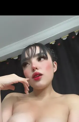 Young woman with bangs and soft makeup posing indoors against a black curtain background.