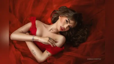 Elegant woman in a red off-the-shoulder dress lying on flowing red fabric in a romantic, moody setting.
