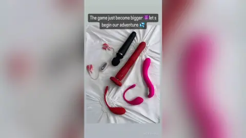 NicoleBella displays a collection of red and black vibrators arranged on a white bedsheet.