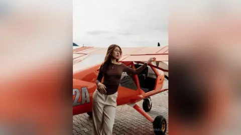 Stylish woman in a brown long-sleeve top and beige trousers posing by a vintage orange airplane under a cloudy sky.