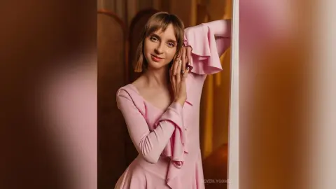 Elegant woman in a pink long-sleeve dress posing gracefully in a warm, soft-lit indoor setting.