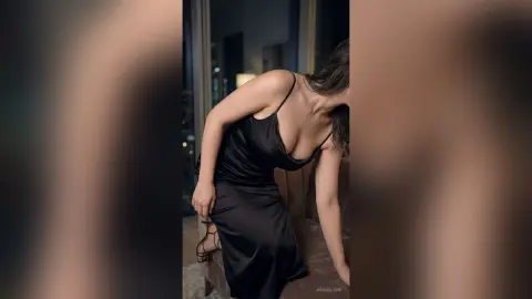 Elegant woman in a sleek black satin slip dress posing in a moody, dimly lit indoor setting.
