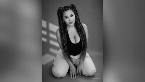 Confident woman with long braids wearing a black bodysuit and white pants in a moody studio setting.