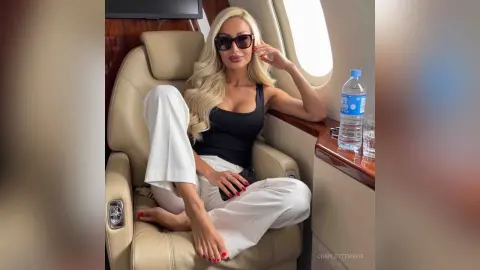 Stylish blonde woman in a black tank top and white trousers relaxing on a luxury private jet seat.