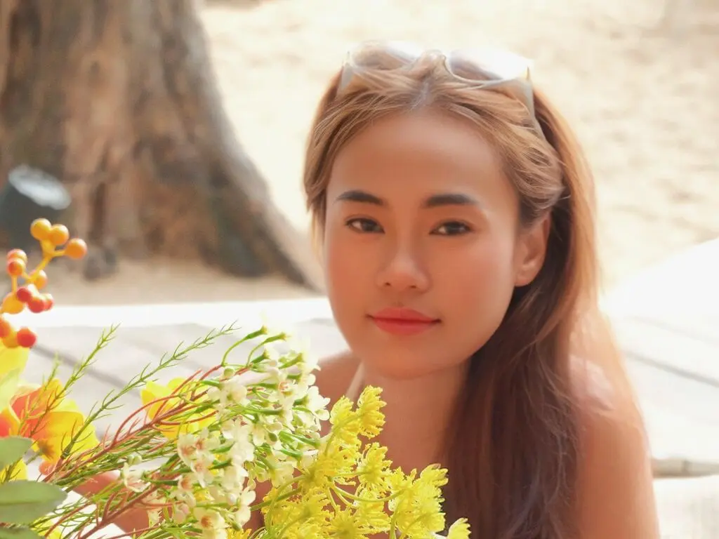 Smiling young woman in casual summer attire relaxing outdoors near bright yellow flowers on a sunny day.